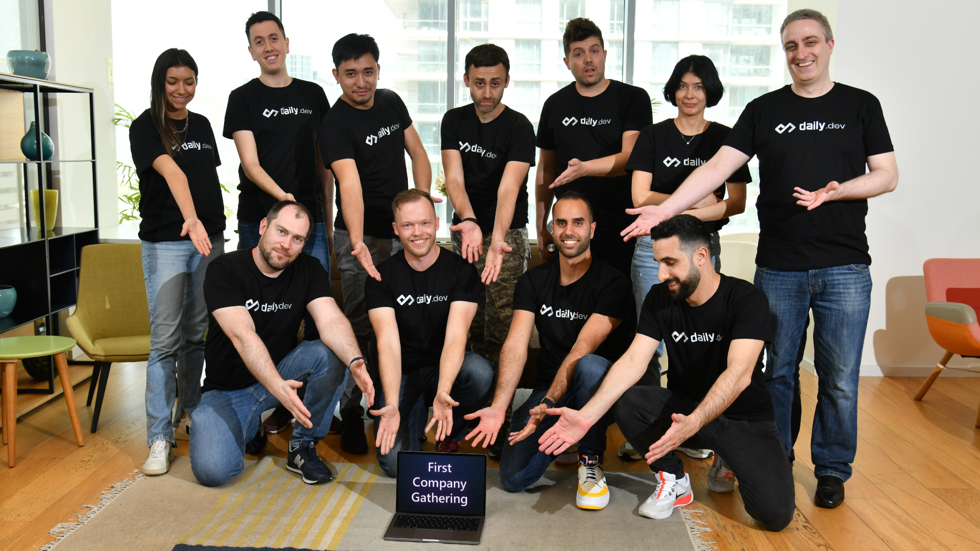 the daily.dev team in branded tshirts pointing at a laptop with the text 'first company gathering' on the screen